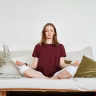 Woman meditating indoors on a sofa, exuding calmness and mindfulness, surrounded by greenery.